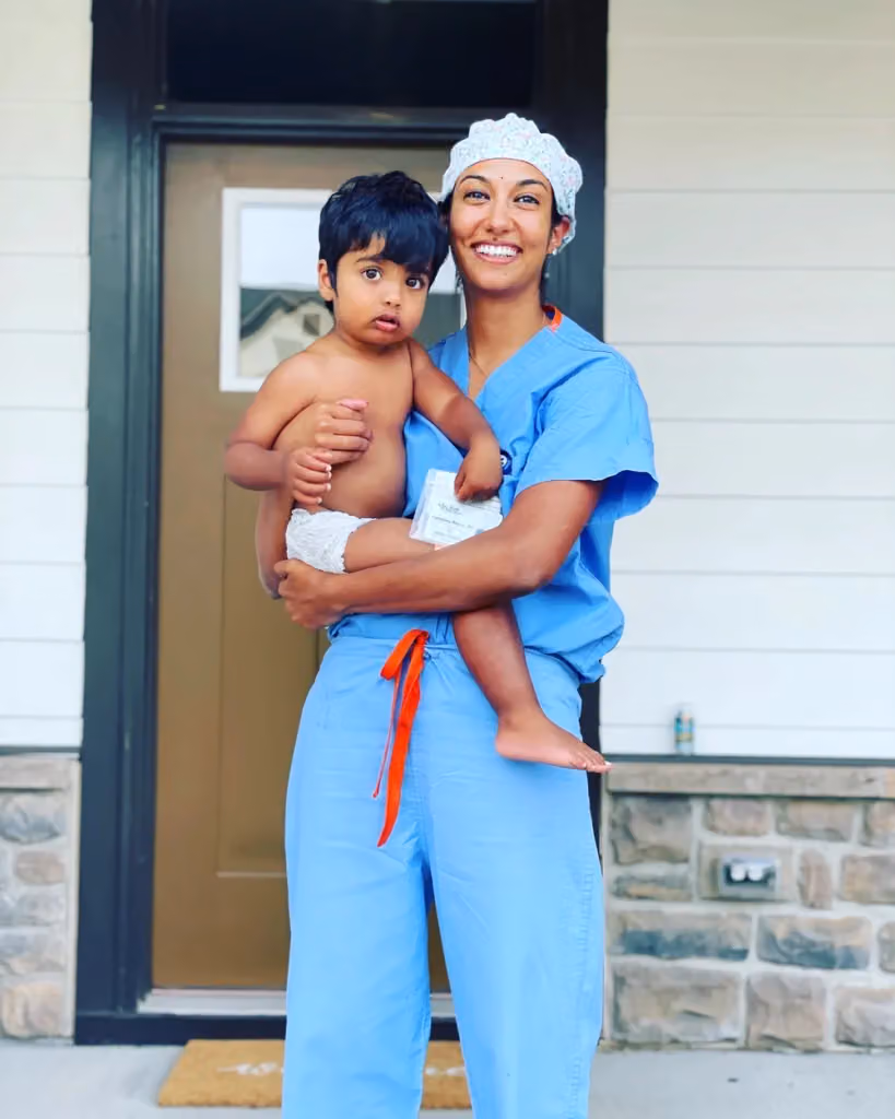 A woman in blue scrubs holding a young child outside a home